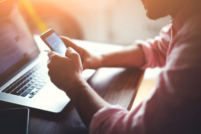 Silhouette of cropped shot of a young man working from home using smart phone and notebook computer