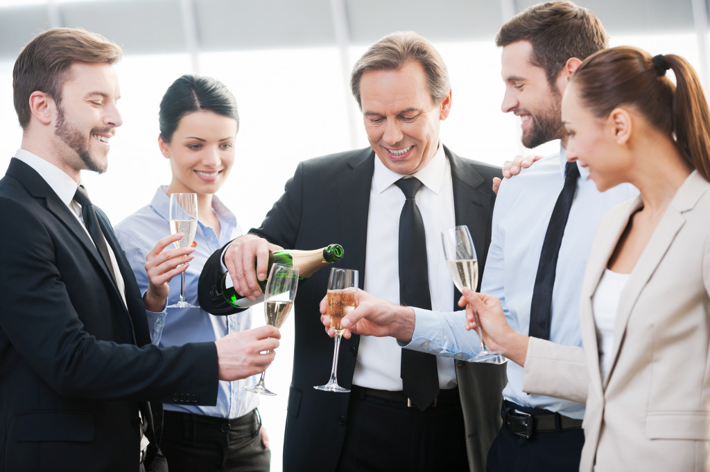 Group of happy business people holding flutes with champagne and smiling while standing close to each other indoors