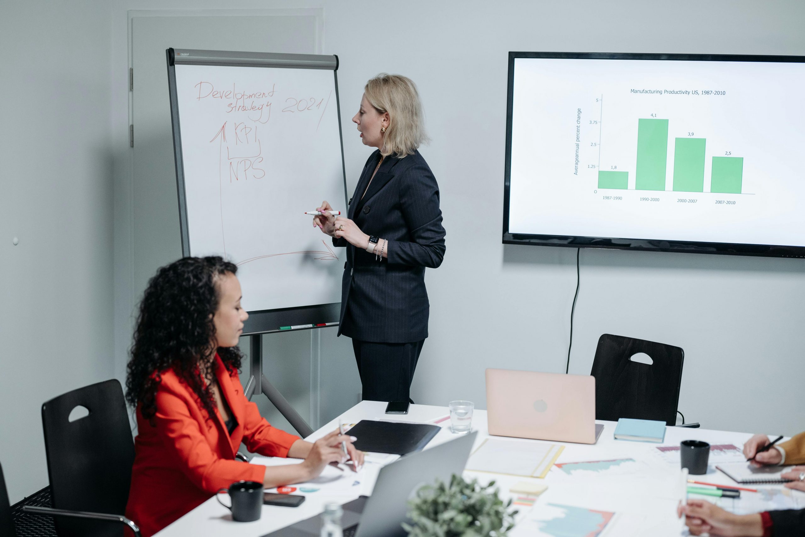 woman in professional attire writing on a whiteboard while presenting to a group of employees