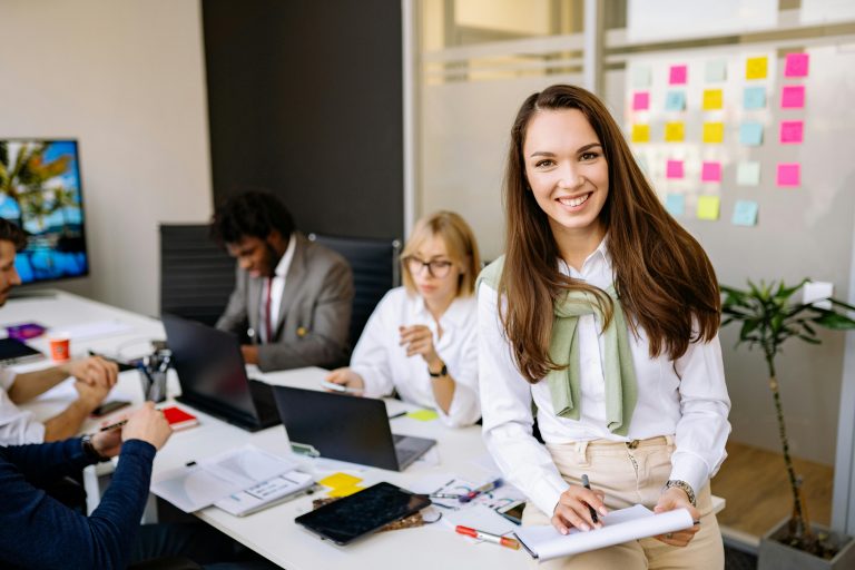 smiling woman in professional attire with colleagues working in the background